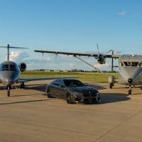 A sleek luxury car and private jets on an airport tarmac under a clear blue sky.